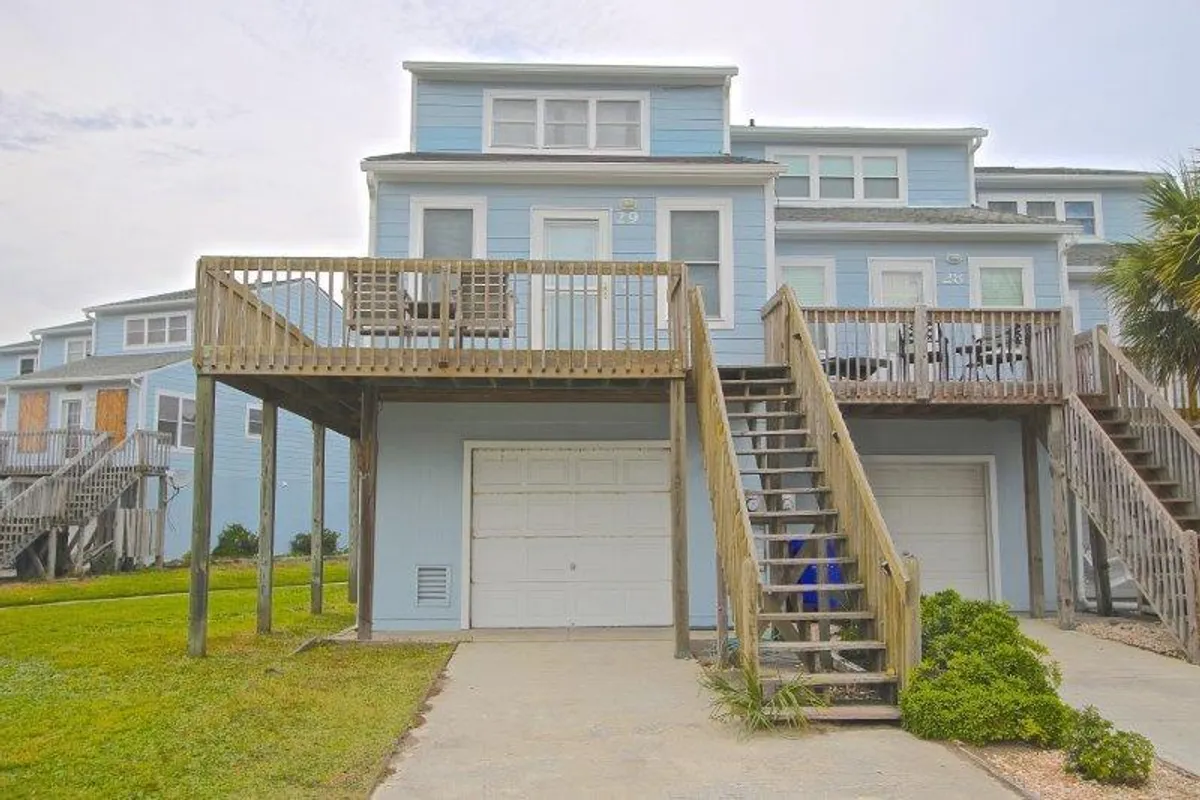 A ground-level exterior shot featuring the private garage with a staircase leading to the main living areas above. The green lawn adds a touch of nature, while the garage door highlights practical storage for beach and vacation gear.