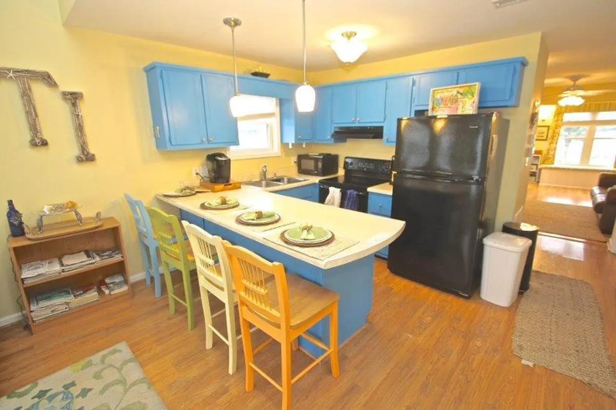 This image showcases a bright kitchen with vivid blue cabinetry, bar stools at the kitchen island, and black appliances. Natural light streams in, illuminating the inviting space designed for cooking, socializing, and shared meals.