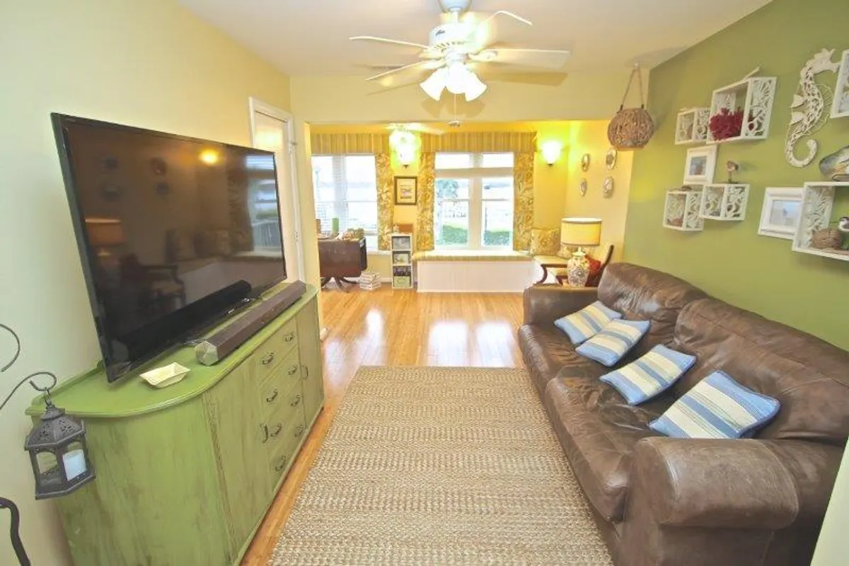 A spacious living area featuring a large TV atop a green dresser, a cozy brown sofa, and windows draped with curtains that let in natural light. The scene exudes comfort for relaxing evenings after a day by the beach.