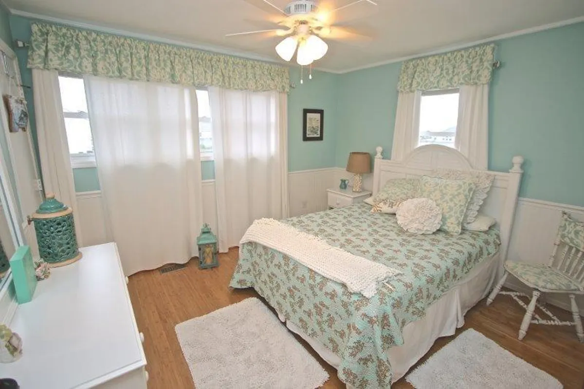 A serene bedroom featuring a bed with patterned bedding, soft curtains, a decorative lamp, and a side table. Morning light streams into the room, enhancing its calm, restful atmosphere.