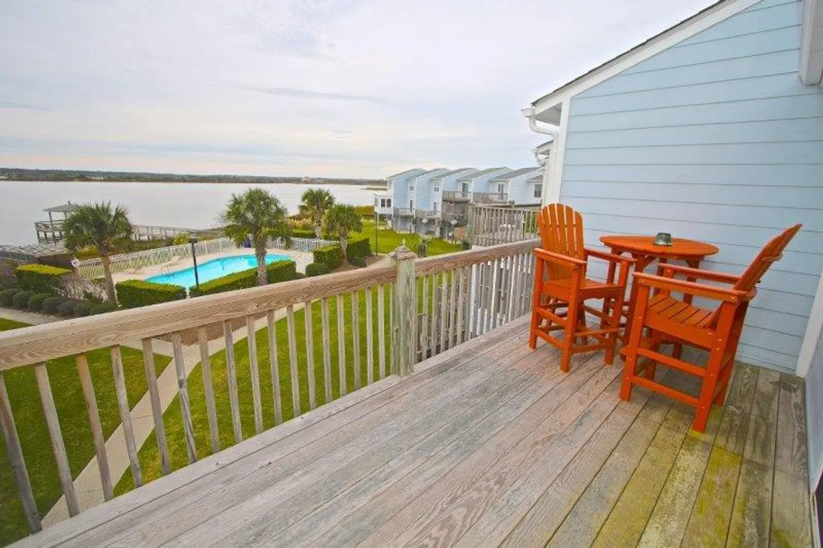 The image features the exterior deck of a charming single-family house at North Topsail Beach. An inviting orange bistro set is staged on the deck, overlooking a sparkling swimming pool framed by lush palm trees. Beyond the pool, a tranquil waterfront view stretches out, perfect for savoring sunsets and serene mornings.