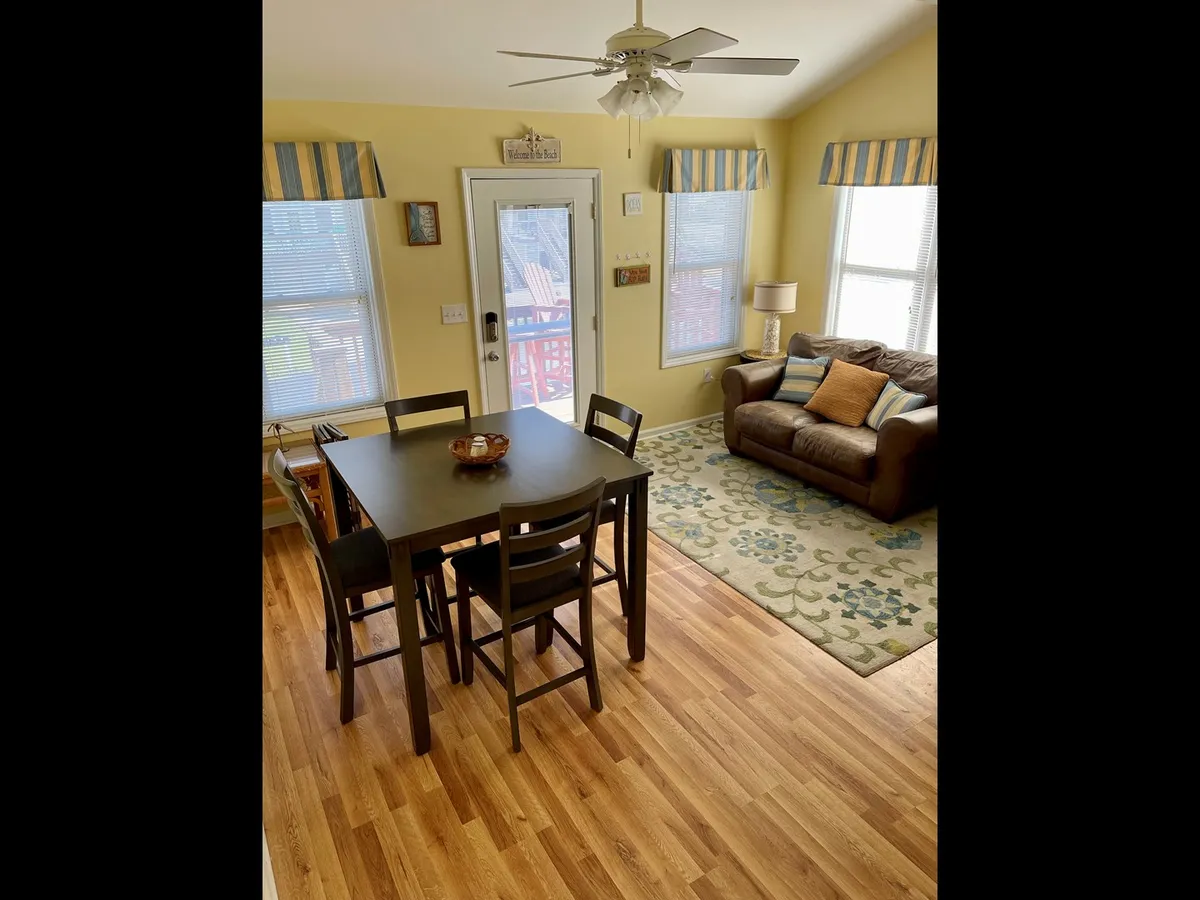 A welcoming view of the dining table adjacent to a cozy living area with a comfortable couch, large windows letting in sunlight, and a decorative rug. The space encourages conversation and relaxation with ample natural light.