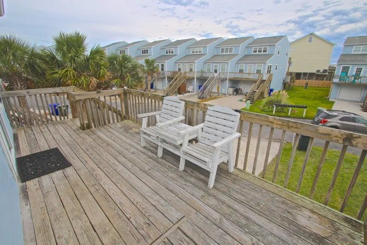 This exterior shot highlights a charming deck outfitted with white wooden chairs and a classic railing, complemented by the backdrop of palm trees and nearby blue houses. The scene invites moments of relaxation, whether for enjoying morning coffee, intimate sunsets, or quiet conversation in the fresh coastal air.