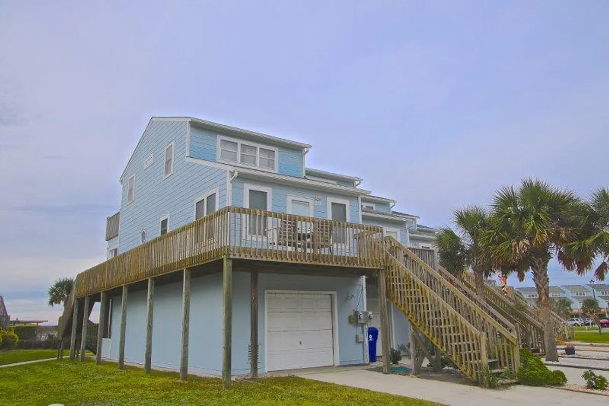 A wooden wraparound deck with palm trees and outdoor seating, attached to a blue house. The deck offers views, areas for relaxation, and easy access between indoor and outdoor spaces, reinforcing seamless transitions for all-day enjoyment.