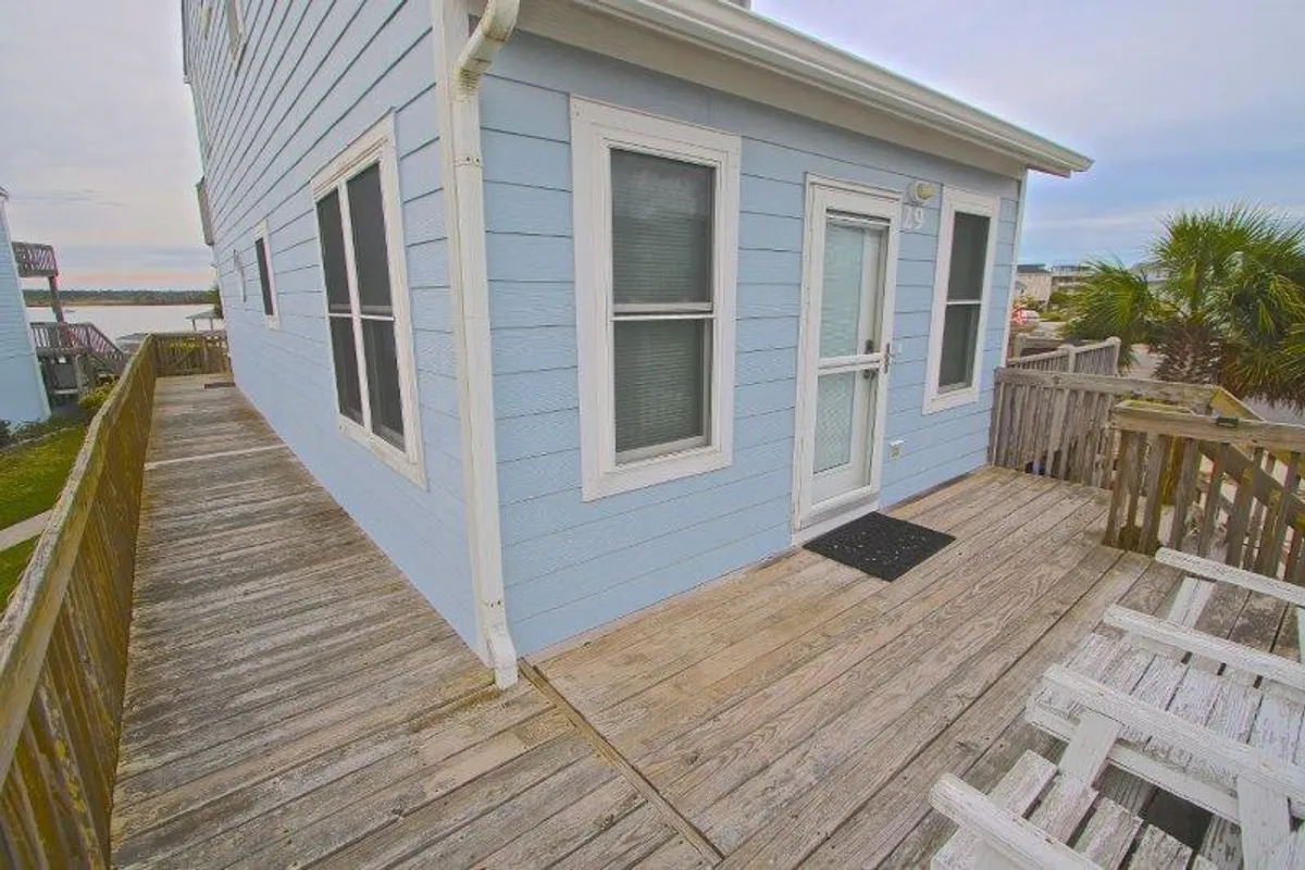 Featuring the light blue exterior of the property, this image includes a wooden deck and an inviting entrance, suggesting a casual, welcoming spot for relaxation or socializing. The deck is an ideal location to watch the sunset or enjoy the gentle coastal breeze.