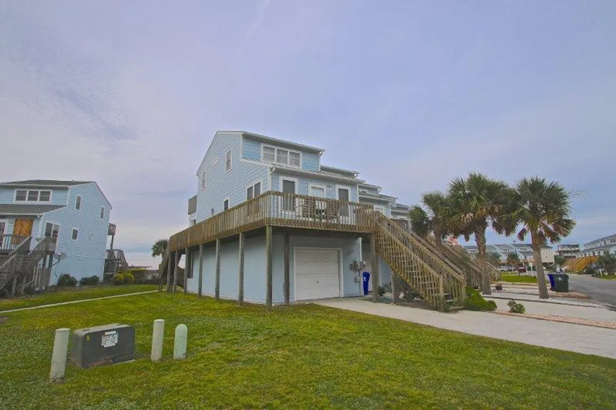 An inviting exterior shot showcasing the house’s elevated deck, accented by palm trees and the blue exterior. The spacious deck is ideal for outdoor meals, games, or simply soaking in views in a casual, relaxed setting.